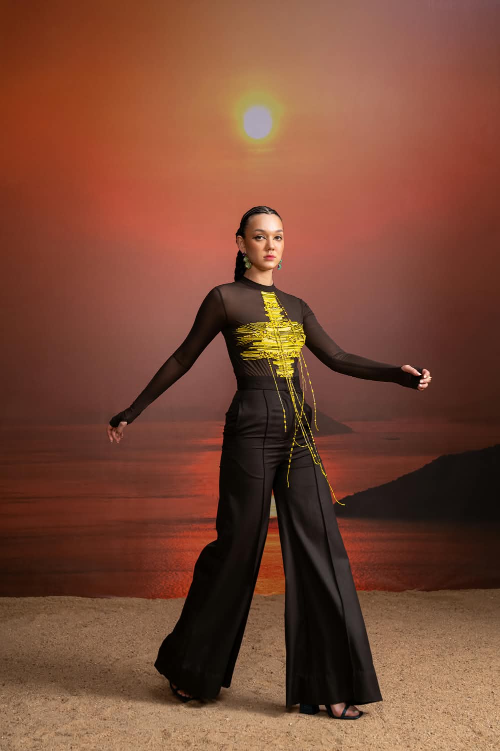 The Solstice ensemble against a burning sky, a sheer mesh top with gold beadwork over wide black trousers.